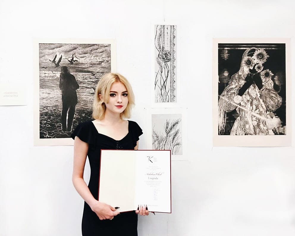 Angelina Ogul na wystawie, z dyplomem, na tle swoich prac graficznych